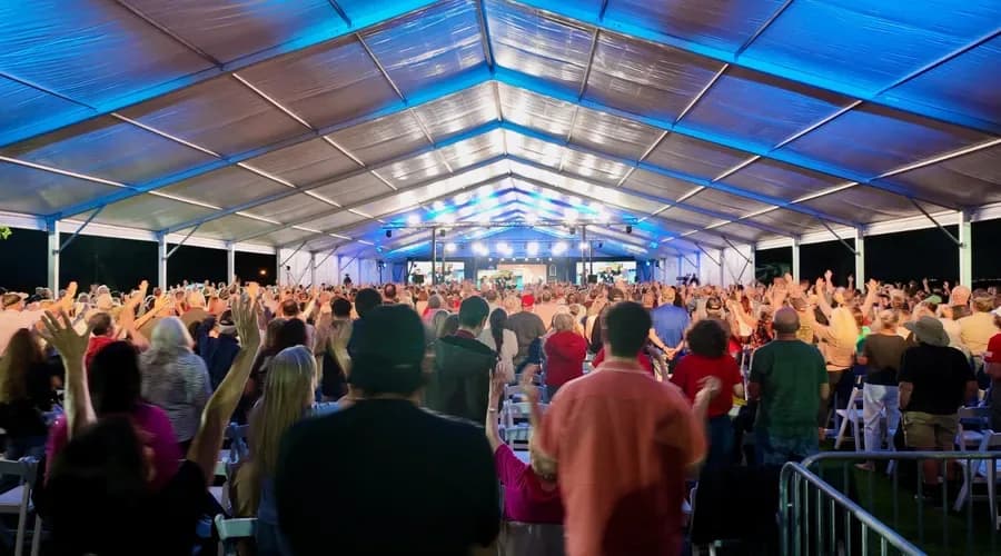 Large crowd gathered under a blue-lit event tent, raising their hands toward a brightly illuminated stage.