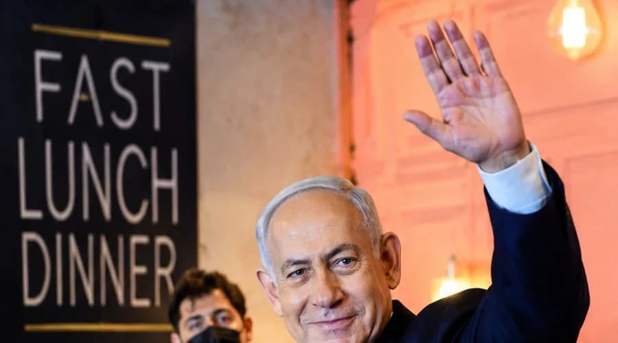 Benjamin Netanyahu waving to attendees at an indoor gathering, surrounded by security personnel and guests.